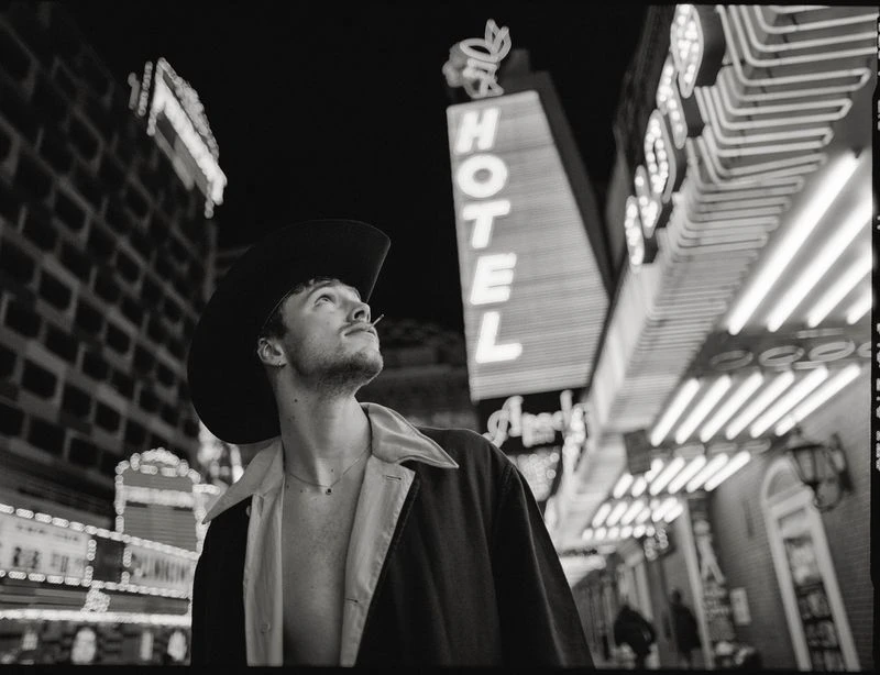 A young adult male in a cowboy hat stands on a city street at night, looking up at illuminated hotel signs and building facades, in black and white.