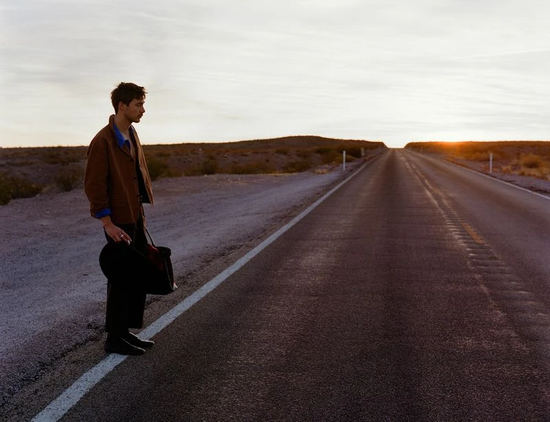 A young man stands on an empty road at sunset, holding a bag and a hat, with a desert landscape stretching into the distance.