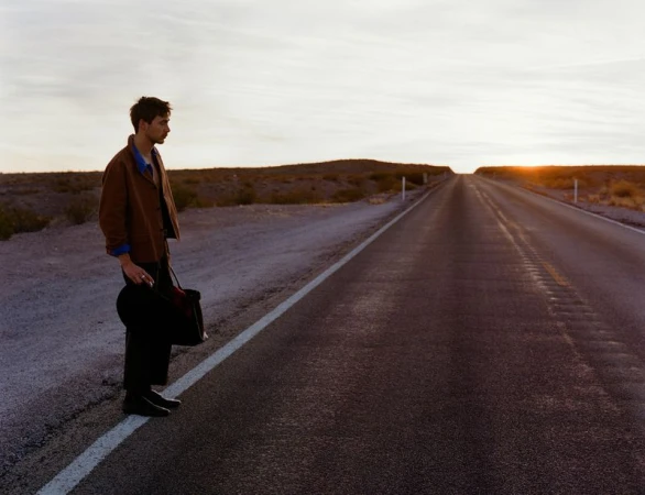 A young man stands on an empty road at sunset, holding a bag and a hat, with a desert landscape stretching into the distance.