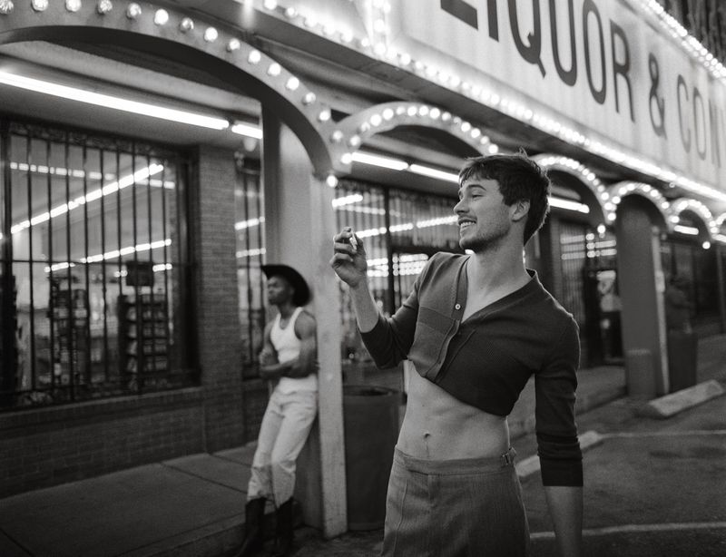 Black-and-white photo of two men outside an illuminated liquor store at night, one wearing a cropped top and the other in cowboy attire.