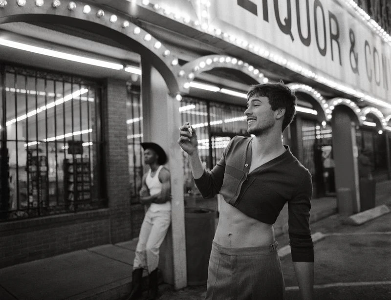 Black-and-white photo of two men outside an illuminated liquor store at night, one wearing a cropped top and the other in cowboy attire.