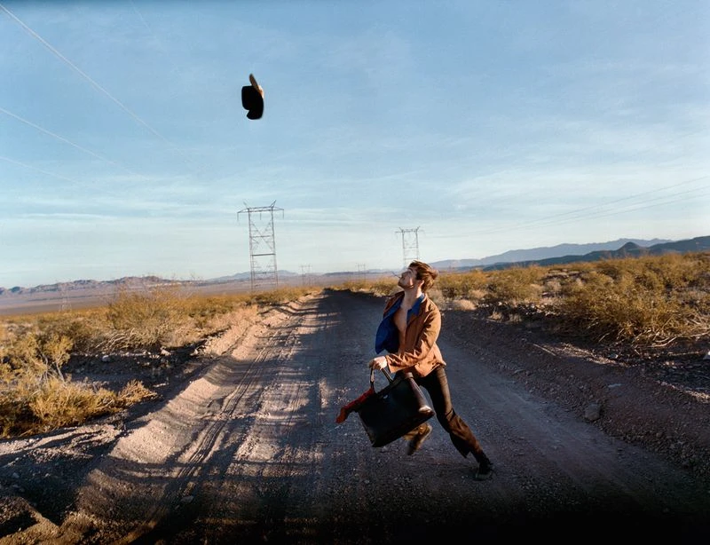 Man walking on dirt road with a purse and jacket flying above in arid landscape on a sunny day, with distant mountains and power lines.