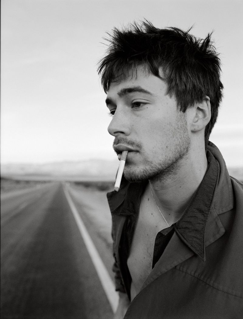 Black and white portrait of a young adult man smoking a cigarette on a desolate desert road with a neutral facial expression.