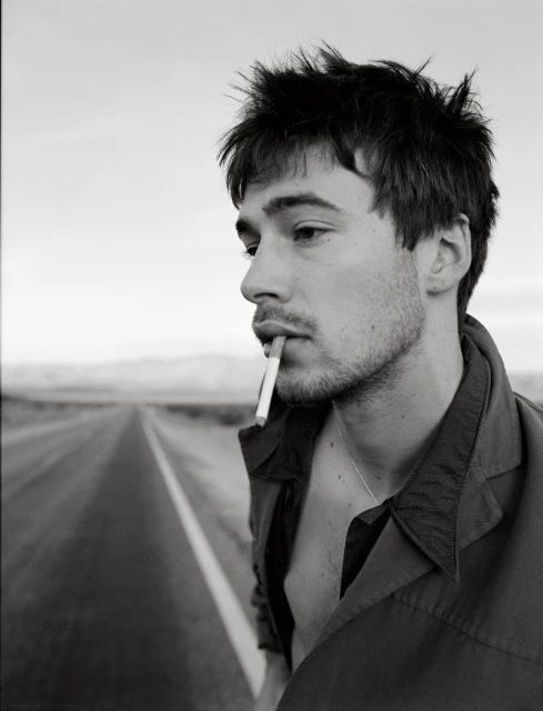 Black and white portrait of a young adult man smoking a cigarette on a desolate desert road with a neutral facial expression.