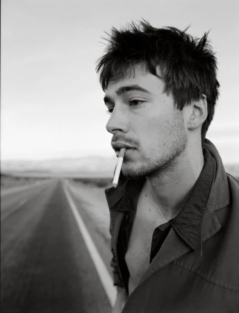 Black and white portrait of a young adult man smoking a cigarette on a desolate desert road with a neutral facial expression.