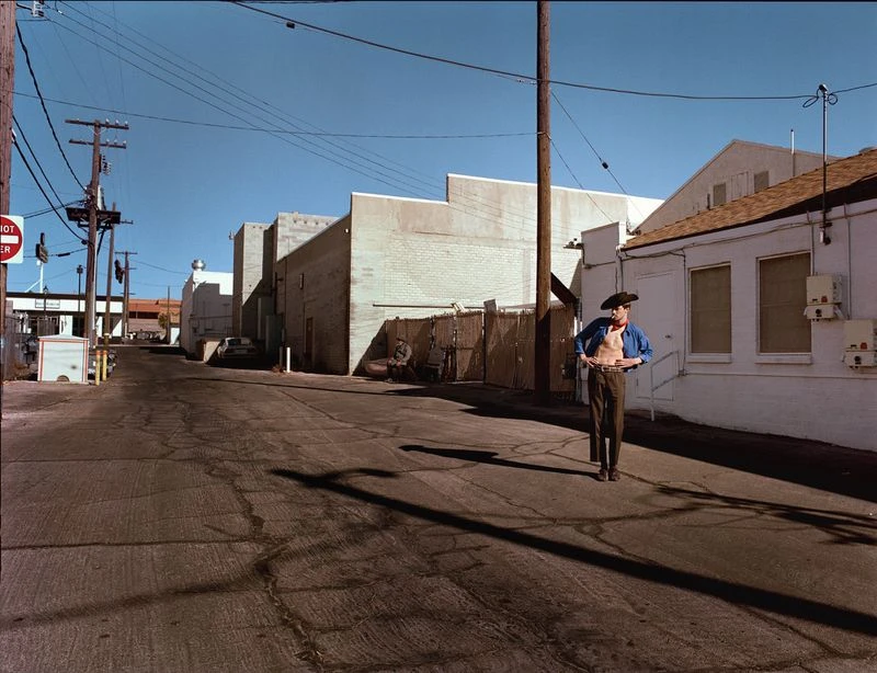 Adult man stands in an urban alleyway wearing a cowboy hat and an open shirt, with buildings and power lines in the background under a blue sky.