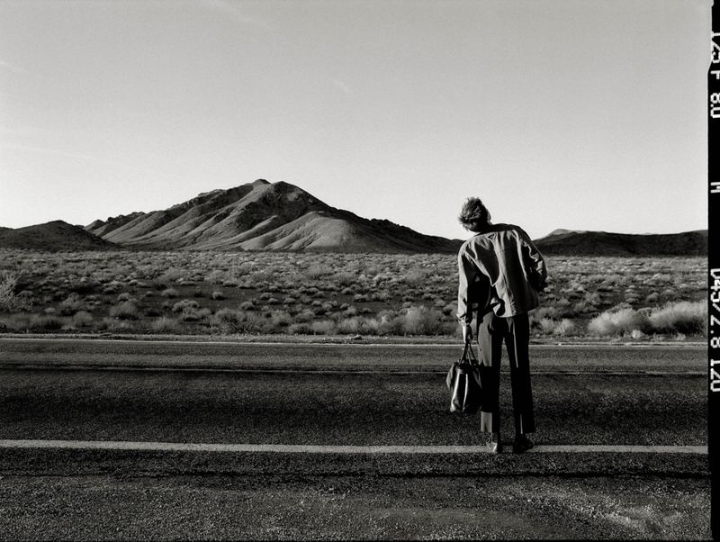 Monochrome shot of an adult standing by a road holding a bag, set against a desert landscape with mountains in the background under a clear sky.