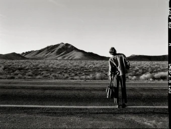 Monochrome image of a person standing by a roadside in a desert landscape holding a bag, shot by Chloe Chippendale for Behind the Blinds.