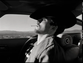 Monochrome portrait of a model wearing a cowboy hat seated inside a car with a desert landscape visible through the window.
