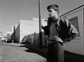 Monochrome image of a young man with a cigarette standing outdoors in an alleyway wearing dark trousers and an unbuttoned shirt.