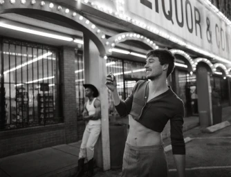 Monochrome street portrait of a smiling young man in a cropped sweater holding a small object outdoors at night.