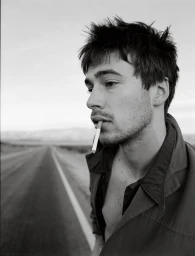 Black and white portrait of a young man holding a cigarette in his mouth while standing beside an empty desert road.