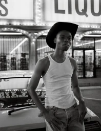 Black man wearing a cowboy hat and white tank top leaning against a vintage car in front of a storefront at night