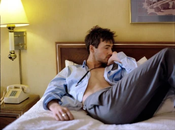 Young man lying on a hotel bed in a partially unbuttoned light blue shirt and grey trousers next to a bedside lamp and phone
