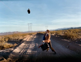 Man leaping on a dirt road in a desert landscape, throwing a hat into the air while holding a black leather bag