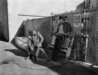Black and white editorial photograph of two men outdoors near a canoe, a chain-link fence, and a California Ave street sign.