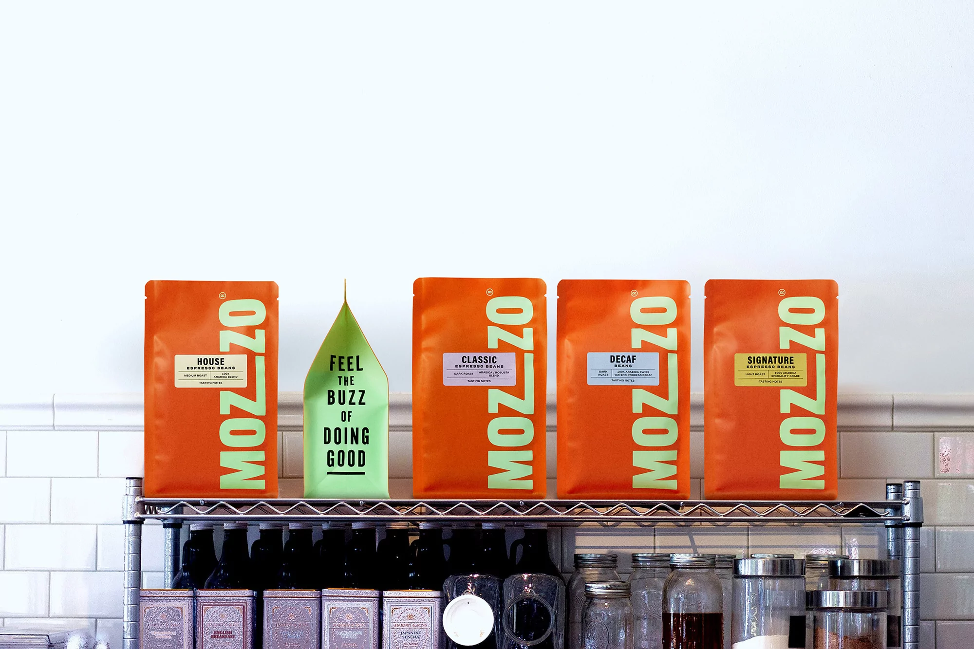 Five Mozzo coffee bean bags are displayed on a metal shelf in a bright setting, including green, orange, and white packaging.