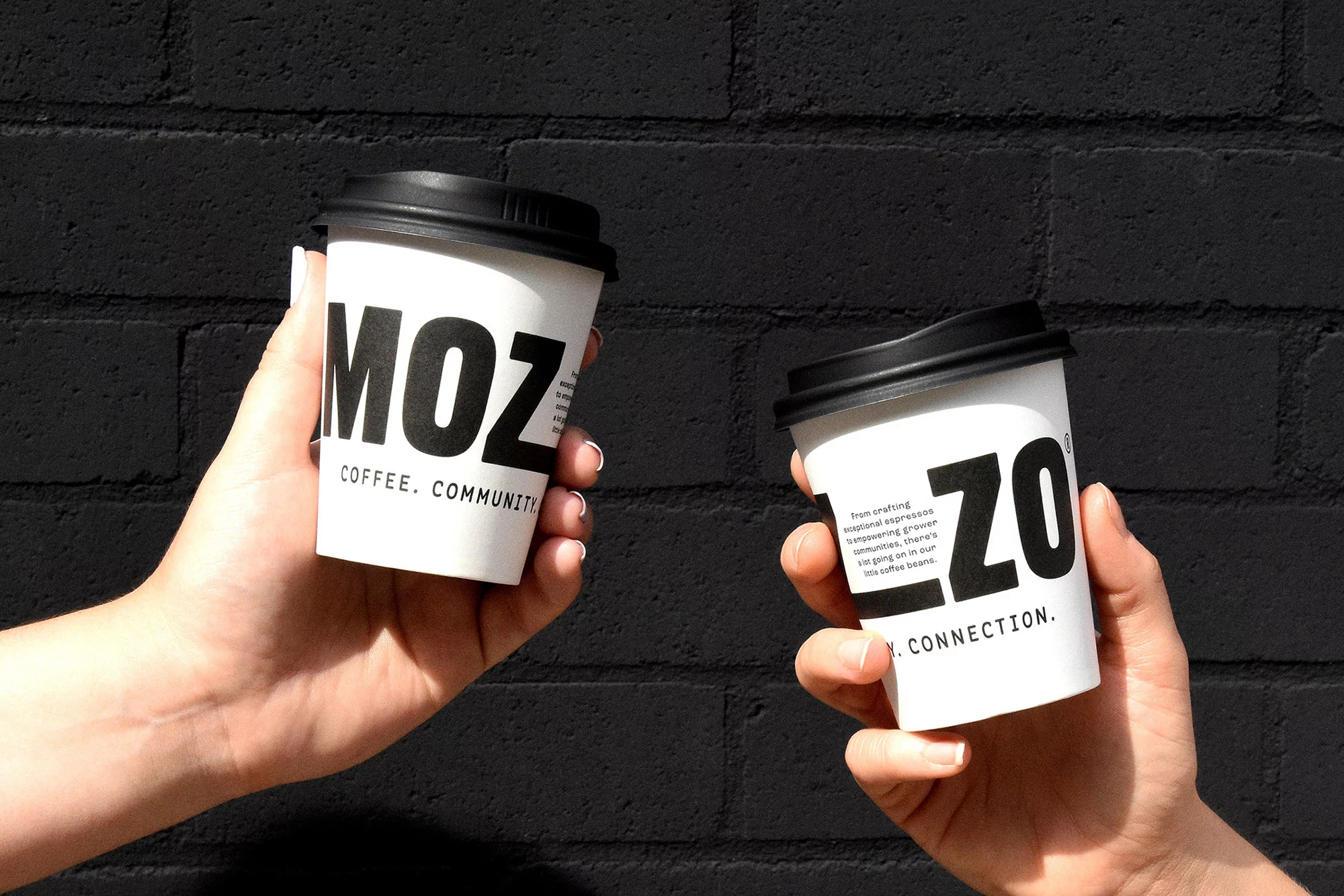 Two hands holding white paper coffee cups with black lids and the word "MOZO" against a dark black brick wall background.