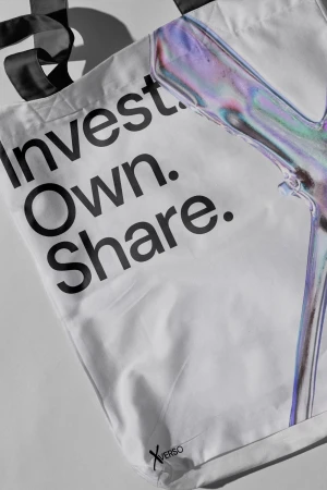 White tote bag featuring bold black typography reading Invest Own Share alongside an iridescent graphic element