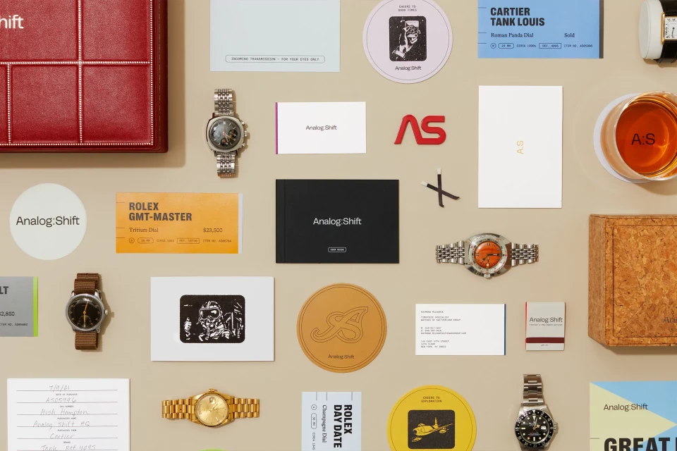 Wristwatches and paper ephemera with the Analog:Shift logo arranged on a neutral background in a high-angle studio shot.