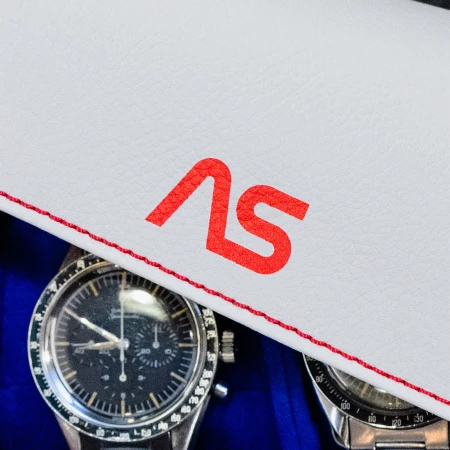 Close-up of two analog wristwatches presented on a white leather surface with a red logo and red stitching, capturing a timeless aesthetic.