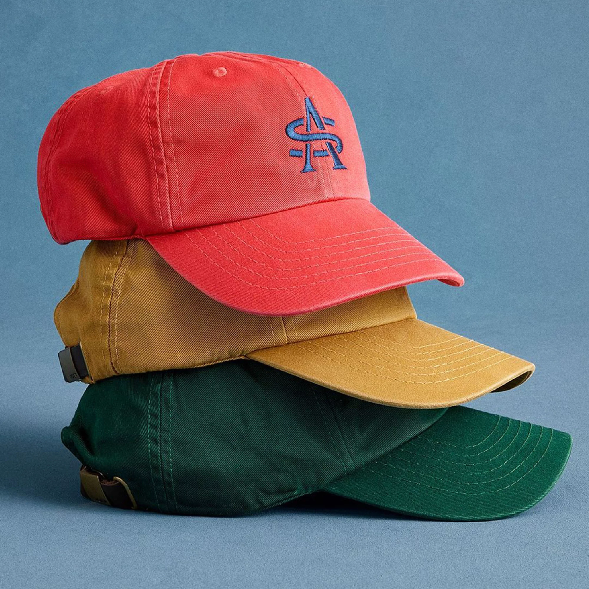 Stack of three baseball caps in red, tan, and green on a light blue background, featuring a simple embroidered monogram logo.