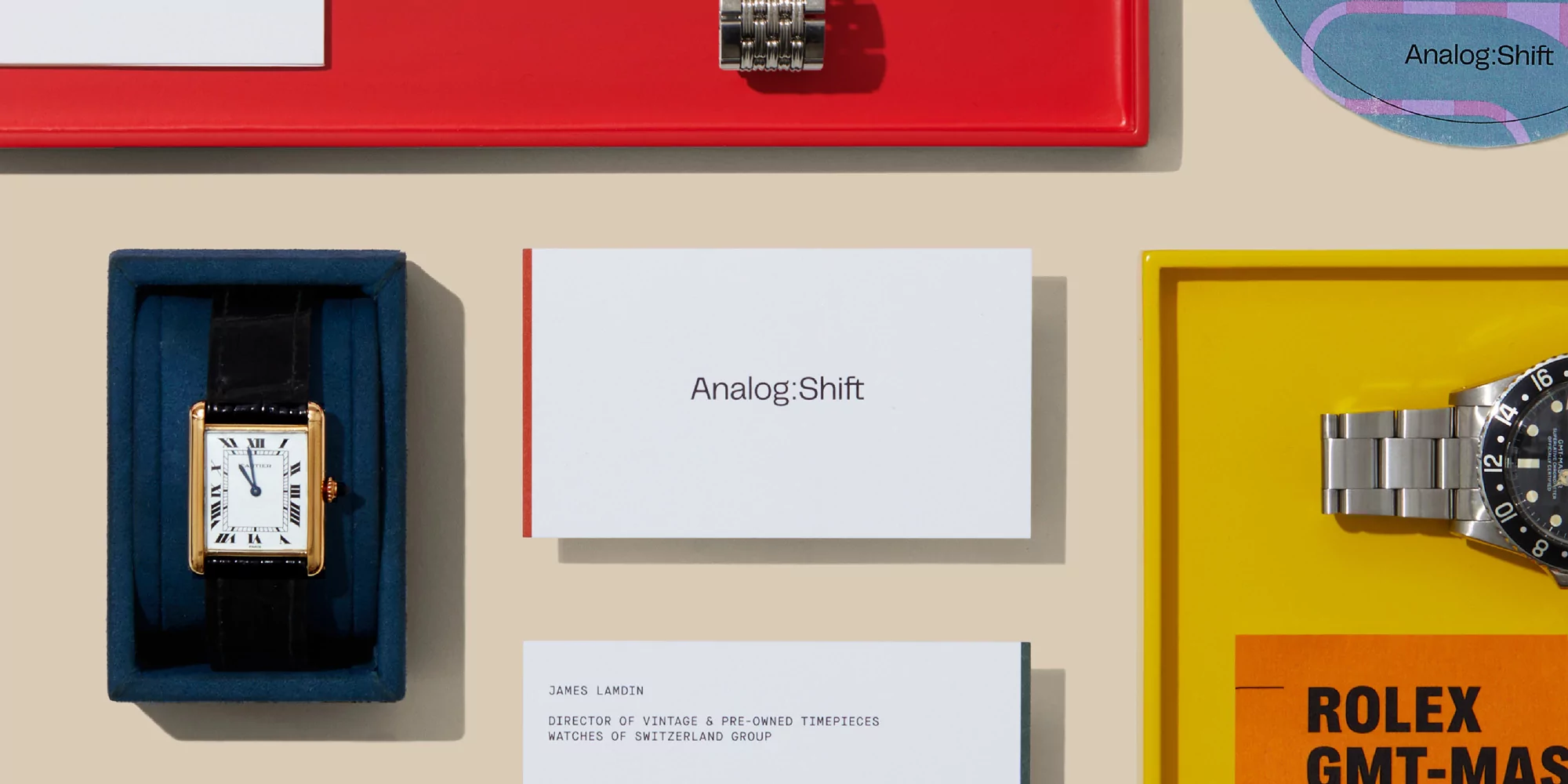 Overhead shot of curated vintage timepieces with business cards on a cream backdrop. Features watches, cards, and trays.