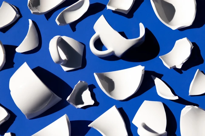 Overhead shot of broken white ceramic mugs and bowls scattered on a vibrant blue surface, creating an abstract, shattered pattern.