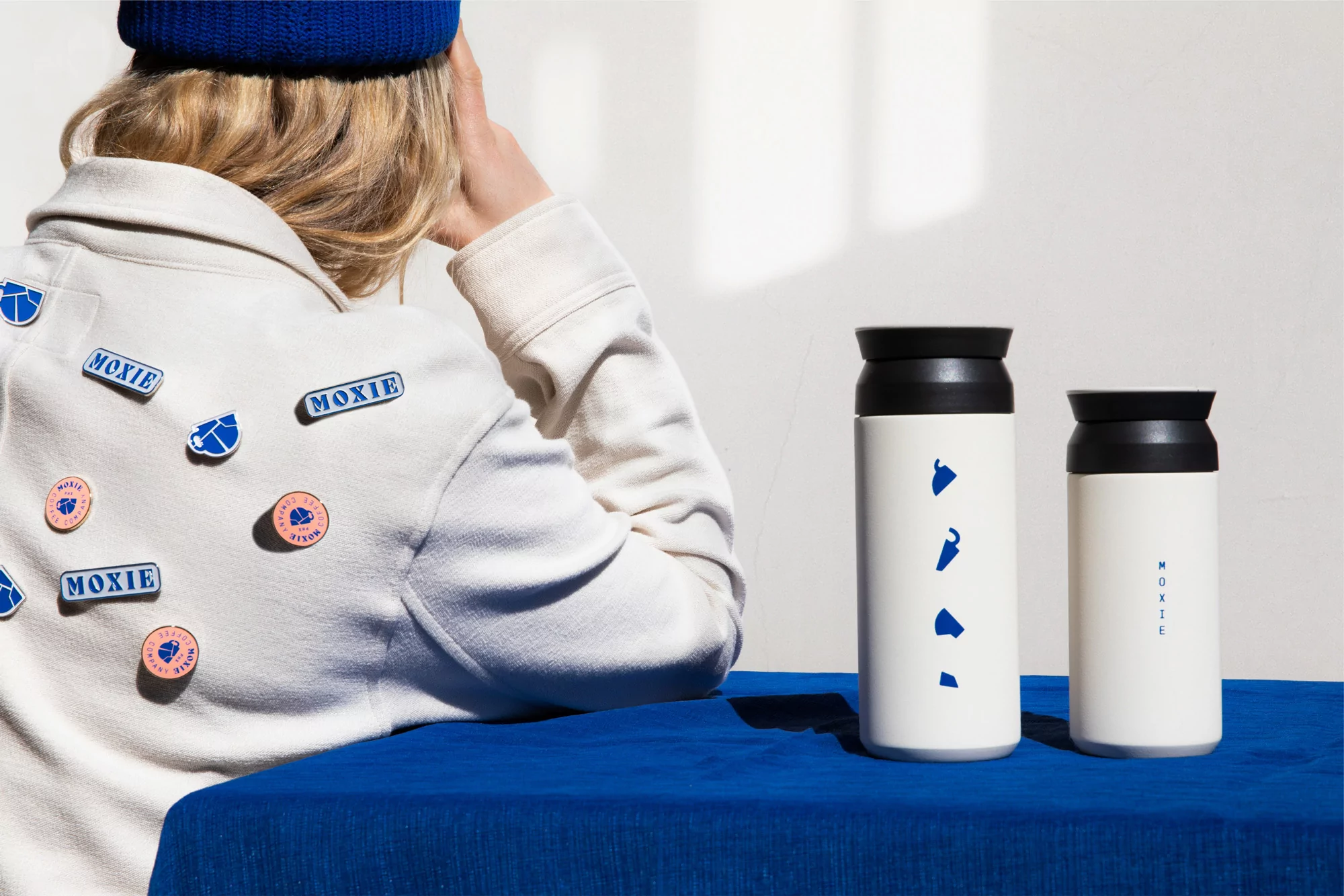 Two white metal bottles with black lids sit on a blue tablecloth with a person wearing a jacket covered in 'Moxie' logo pins.