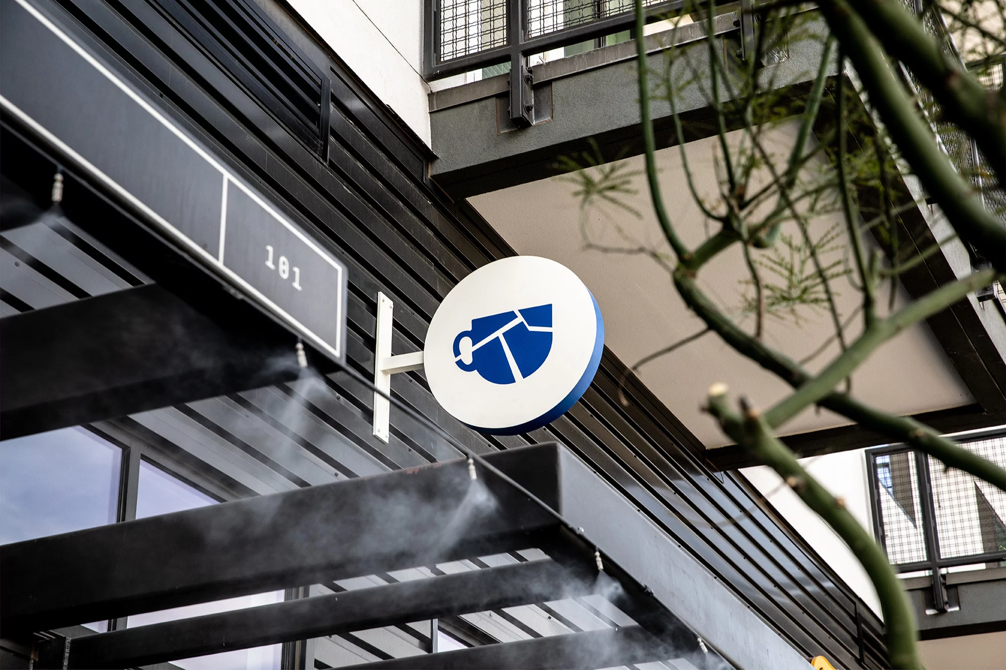 Exterior view of a building with a blue and white coffee cup logo sign and '101' street number under a dark awning.