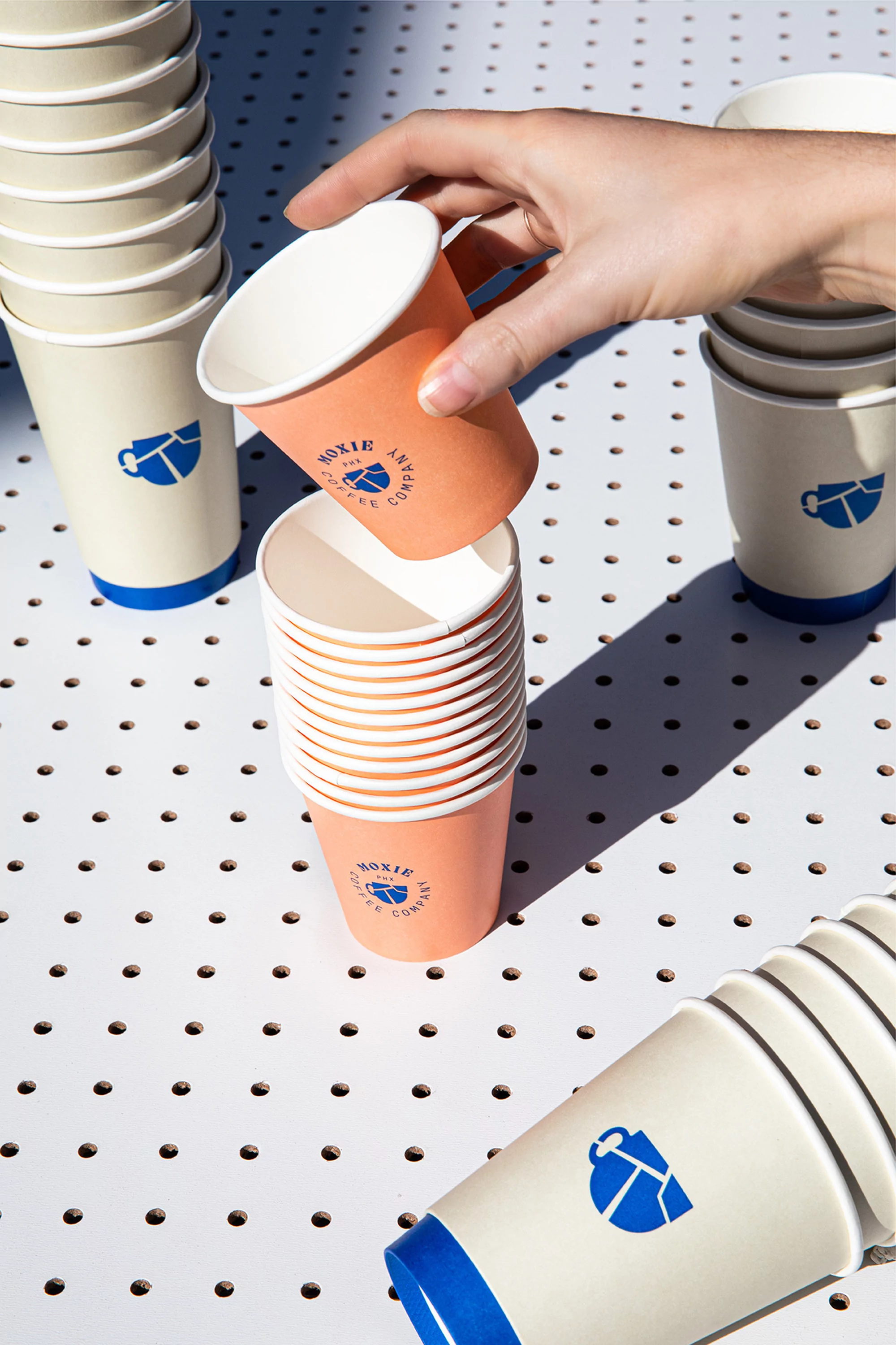 Hand stacking paper coffee cups branded with logo on a white perforated table with natural lighting and visible shadows. Product photography.