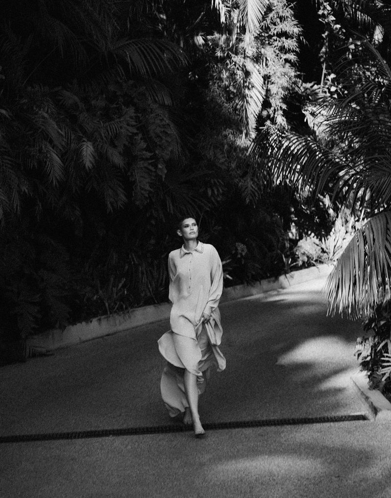 Adult woman walking barefoot in long collared dress with palm trees lining the road in black and white, outdoor, full shot.