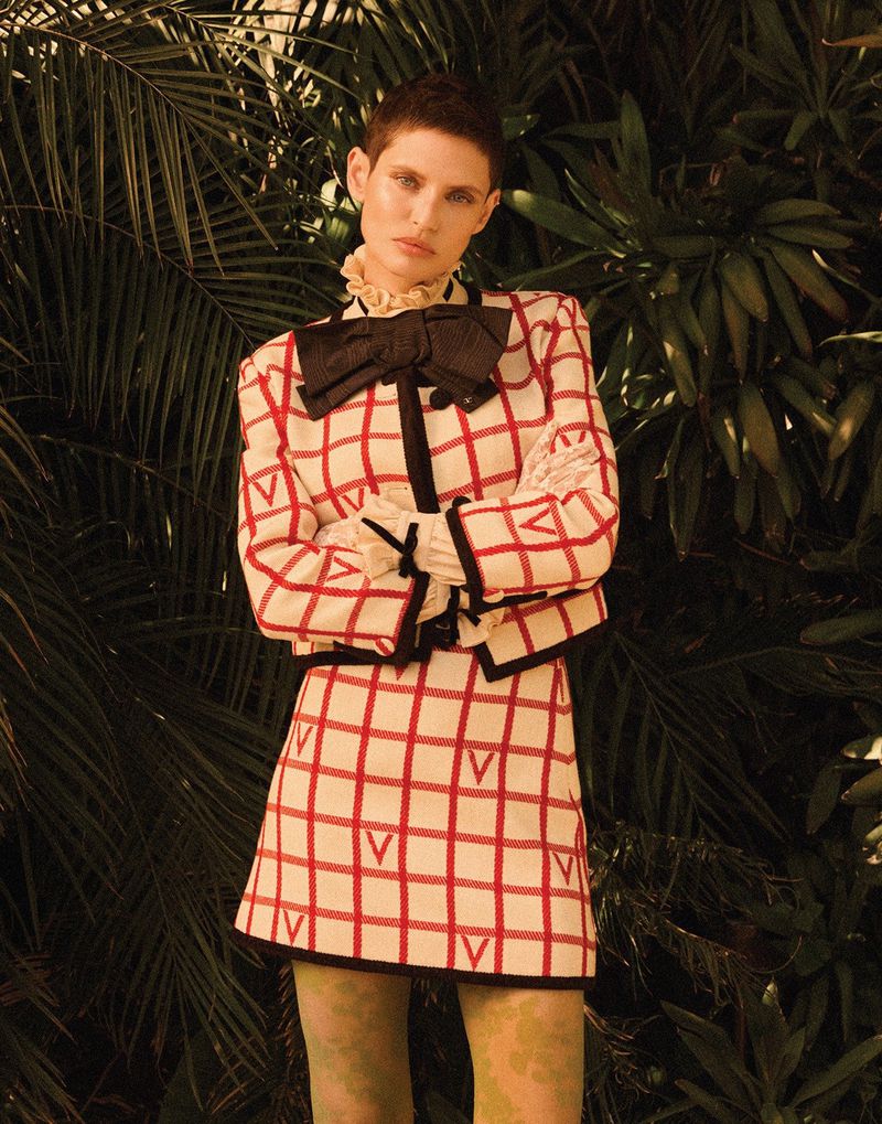 A woman poses in a red and white checkered skirt suit with a black bow, standing against a lush green tropical plant background.