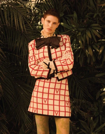 A woman poses in a red and white checkered skirt suit with a black bow, standing against a lush green tropical plant background.