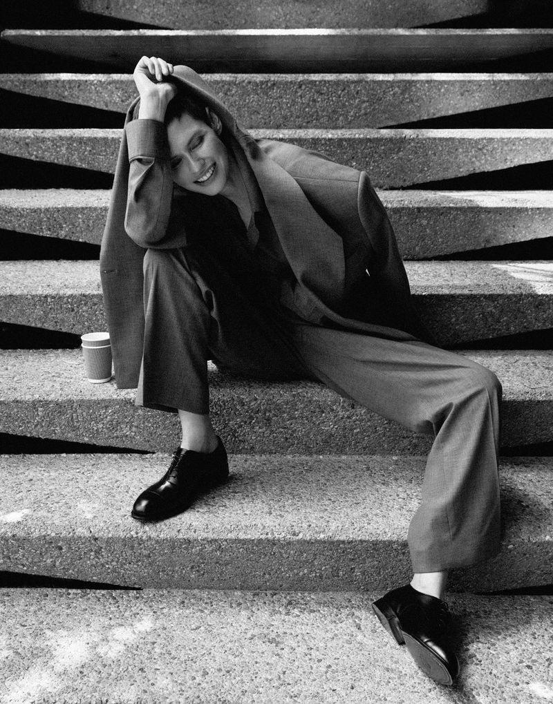 A woman in a suit sits on concrete steps in a monochromatic fashion shot with hard lighting, accented by black dress shoes and nearby cup.