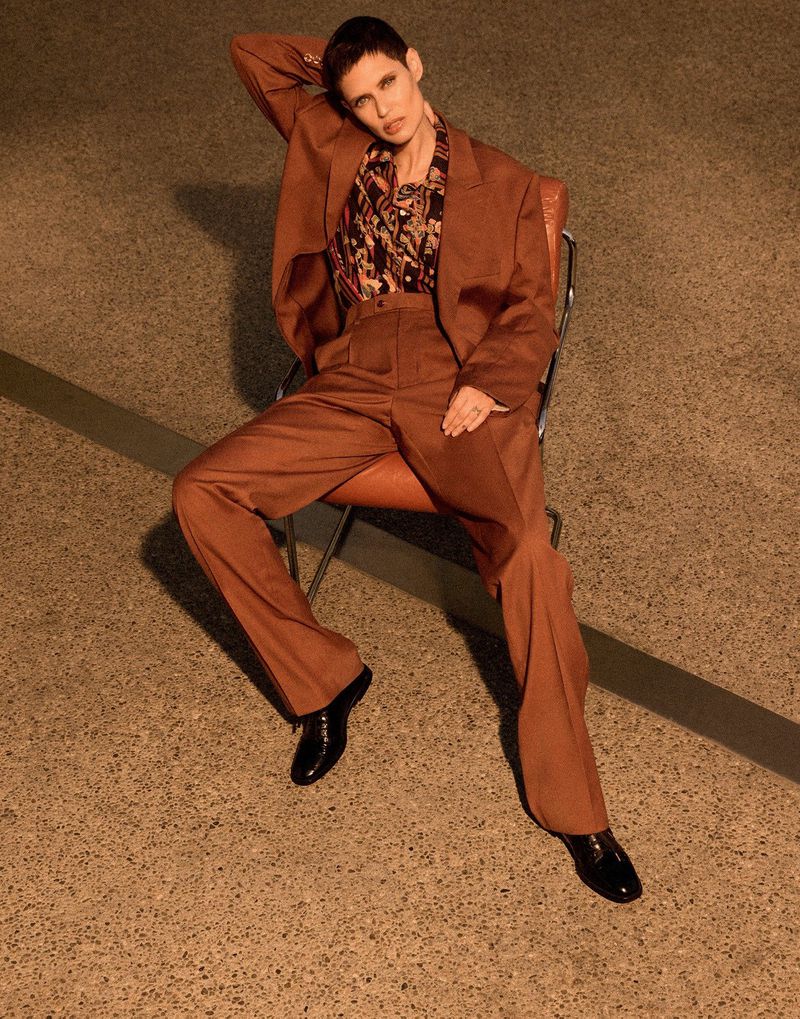 Woman in a brown suit sitting on a chair. She is wearing black shoes and has short, dark hair in an indoor setting with a tan floor.