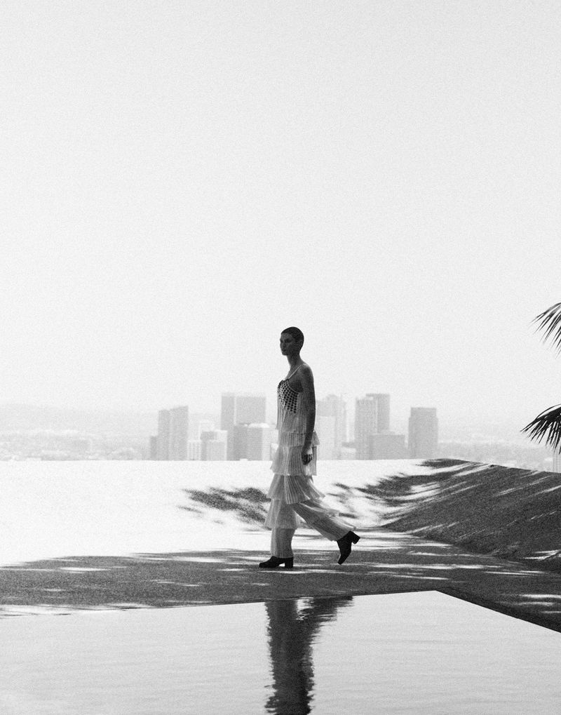 A woman in a layered dress walks by a pool overlooking an urban skyline on a bright day, reflecting in the water.