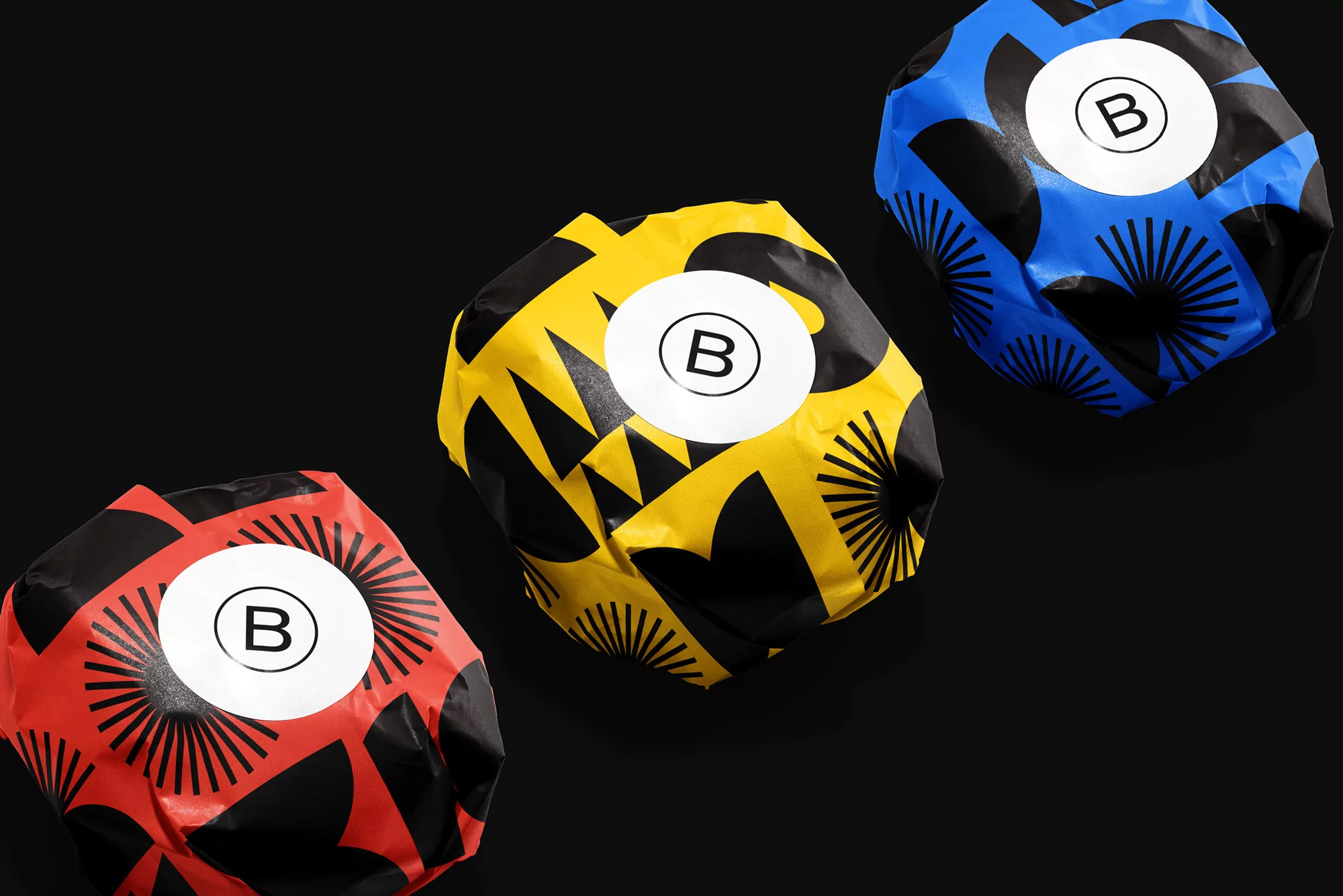 Abstract product shot featuring three colorful packages with black geometric patterns and white circular logos against a black background.