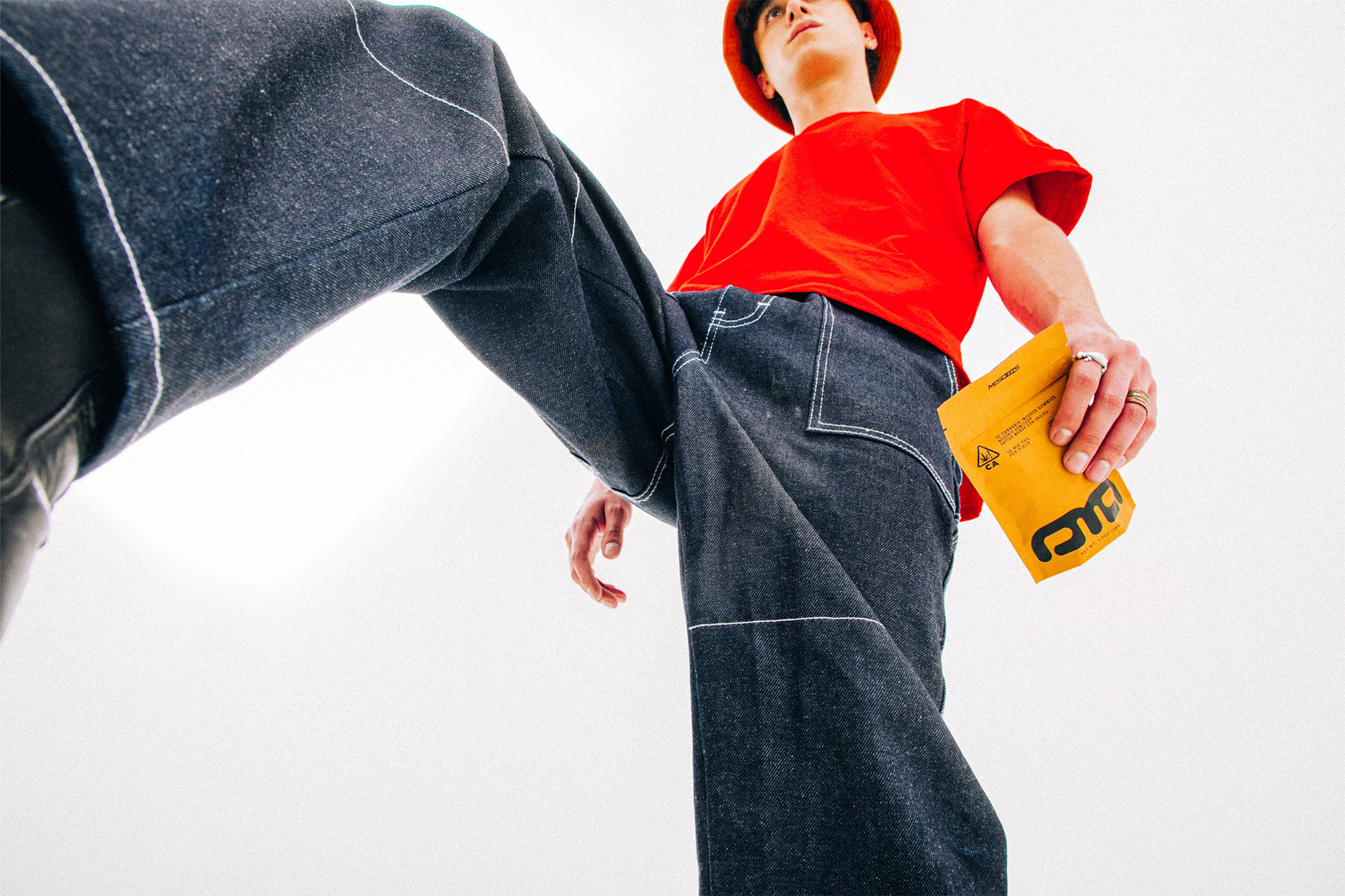 Low-angle fashion shot of a young adult in a red t-shirt, blue jeans, and an orange hat holding a yellow package in a white environment.