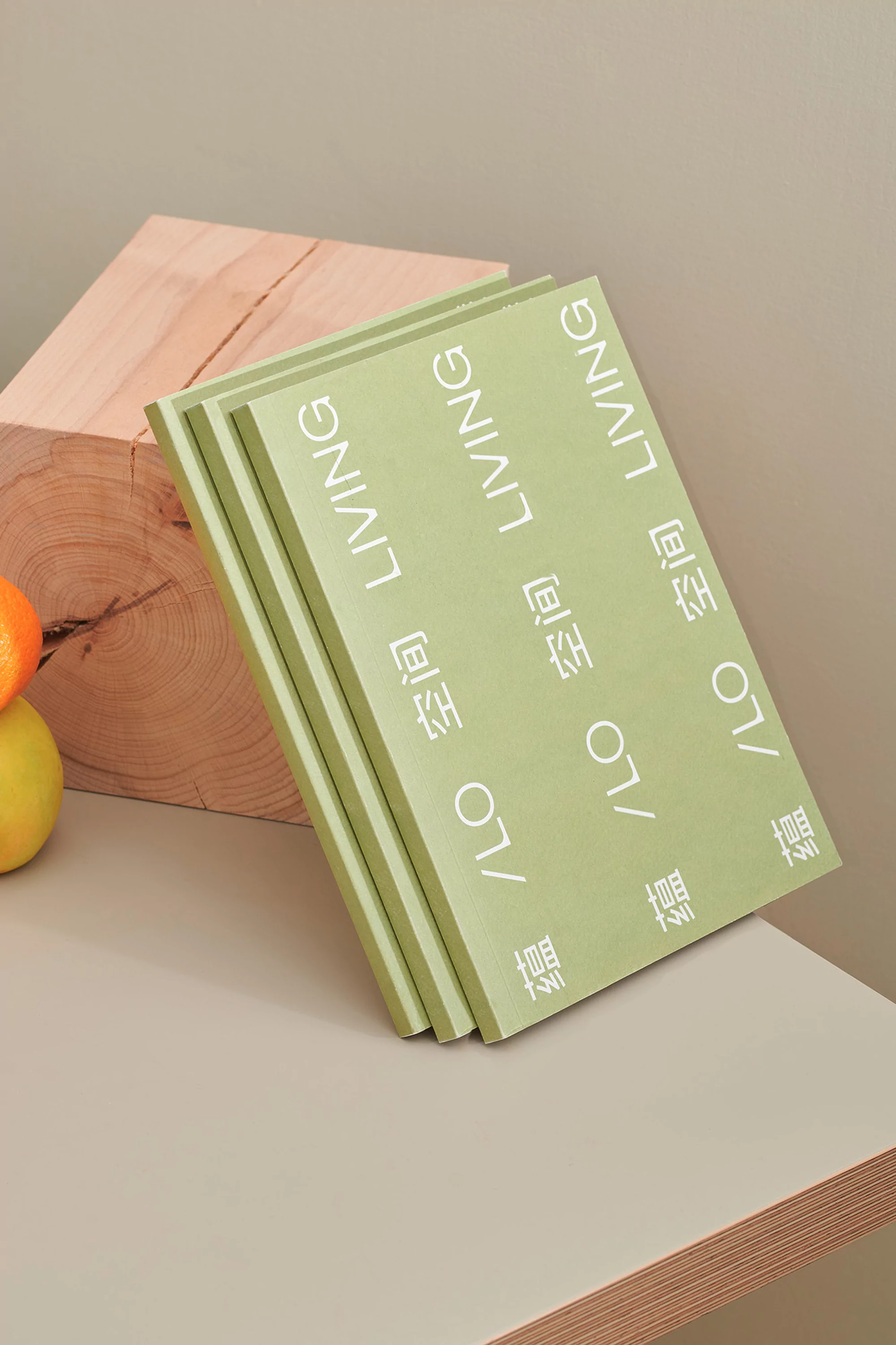 Stack of light green magazines with the words 'LO LIVING' in white, sitting on a beige surface with a wooden block and fruit.