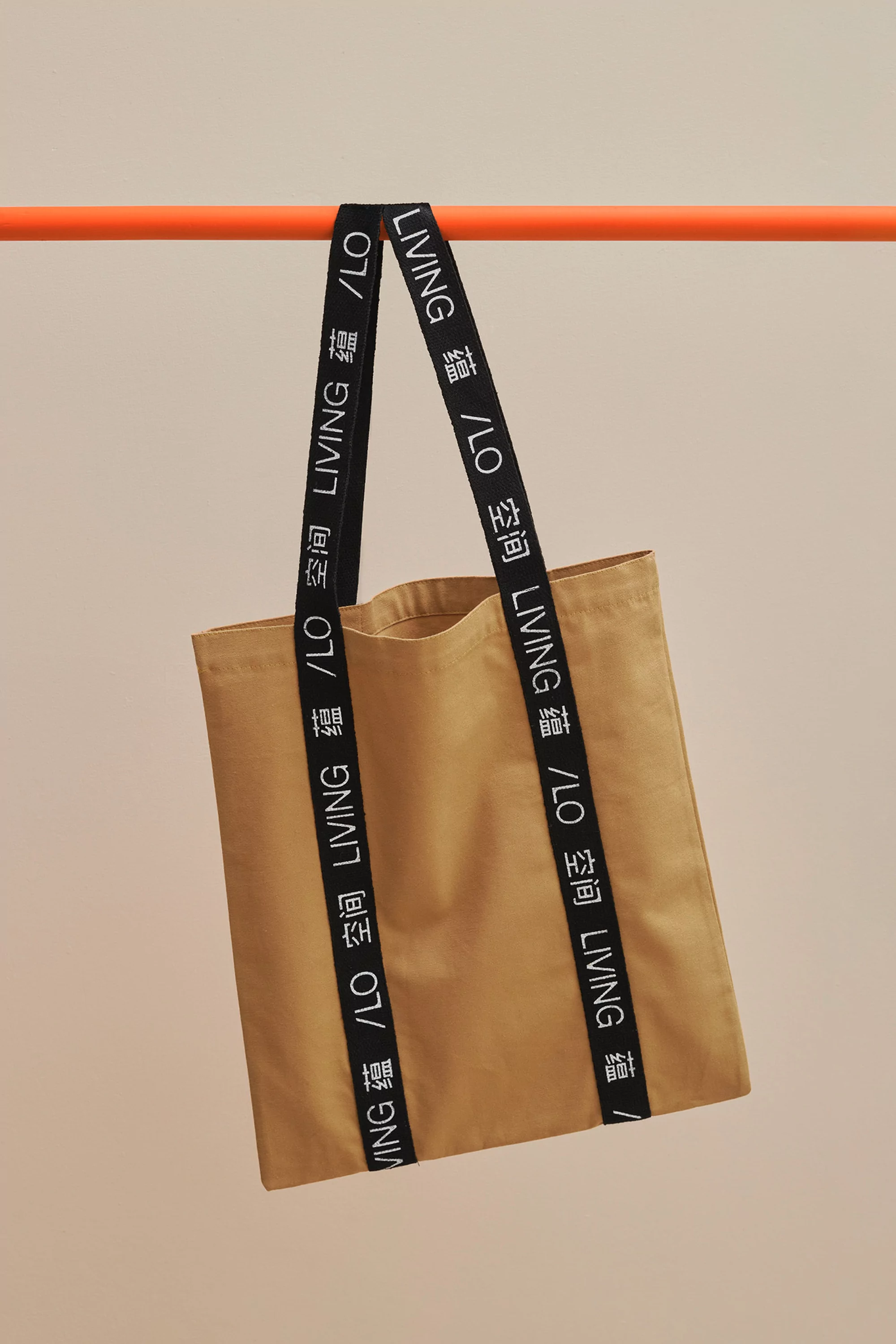 Tan-colored tote bag with black straps featuring white lettering, hanging from an orange bar against a neutral background.