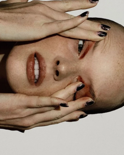 Close-up portrait of a fair-skinned woman with dark manicure covering part of her face with her hands on a white backdrop.