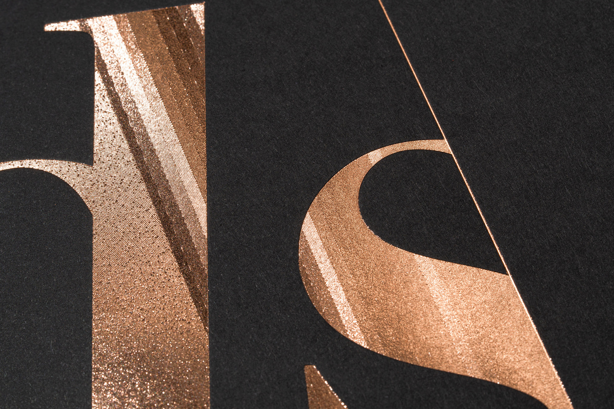 Textured paper with metallic rose-gold lettering featuring oversized, abstracted letterforms against a matte black background, closeup.