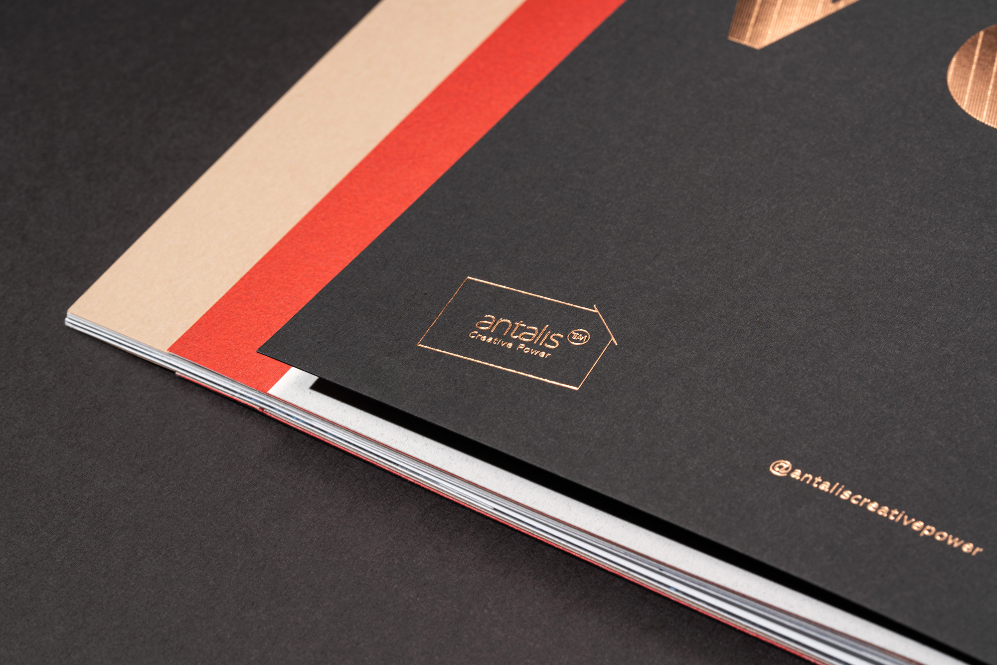 Stack of offset-printed brochures featuring a black cover with bronze foil stamping of the brand logo and marketing hashtag.