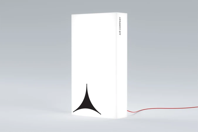 A rectangular white lamp with a black geometric logo and the text AIR COMPANY, displayed against a grey background, lit by soft light.
