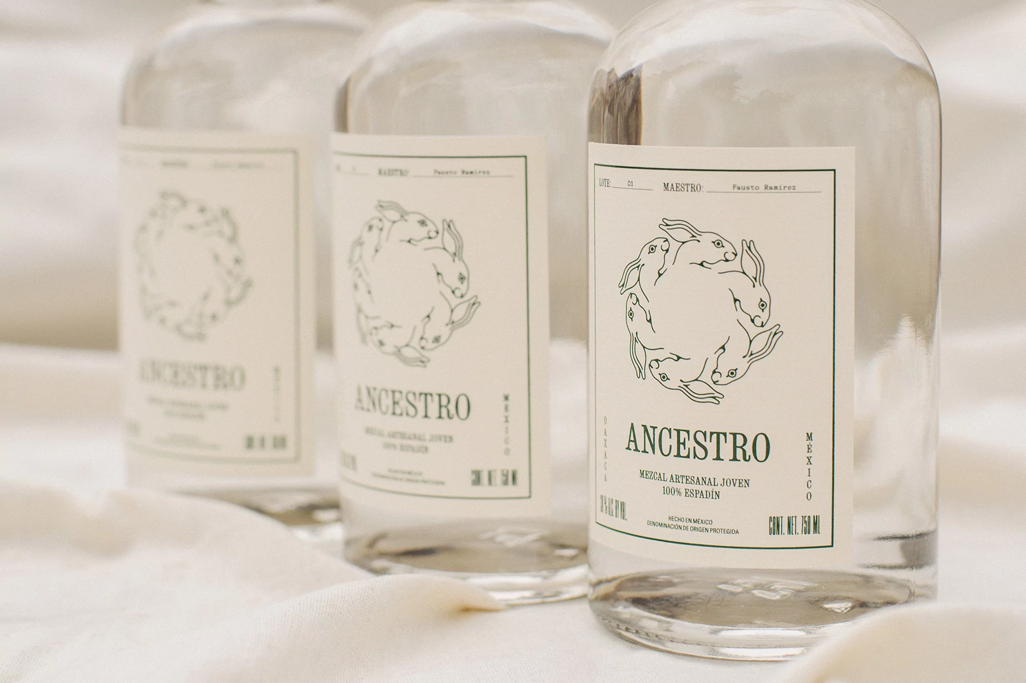 Three clear glass bottles of Ancestro Mezcal are displayed on a white cloth, labels featuring rabbits, creating a clean product shot.