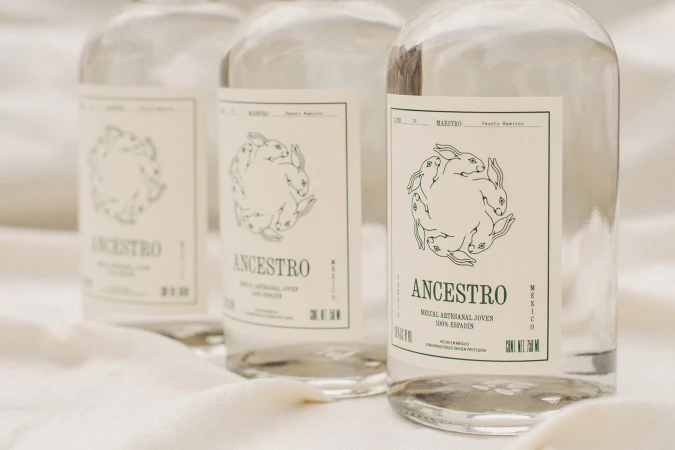 Three clear glass bottles of Ancestro mezcal with white labels featuring rabbit illustrations resting on soft cream fabric