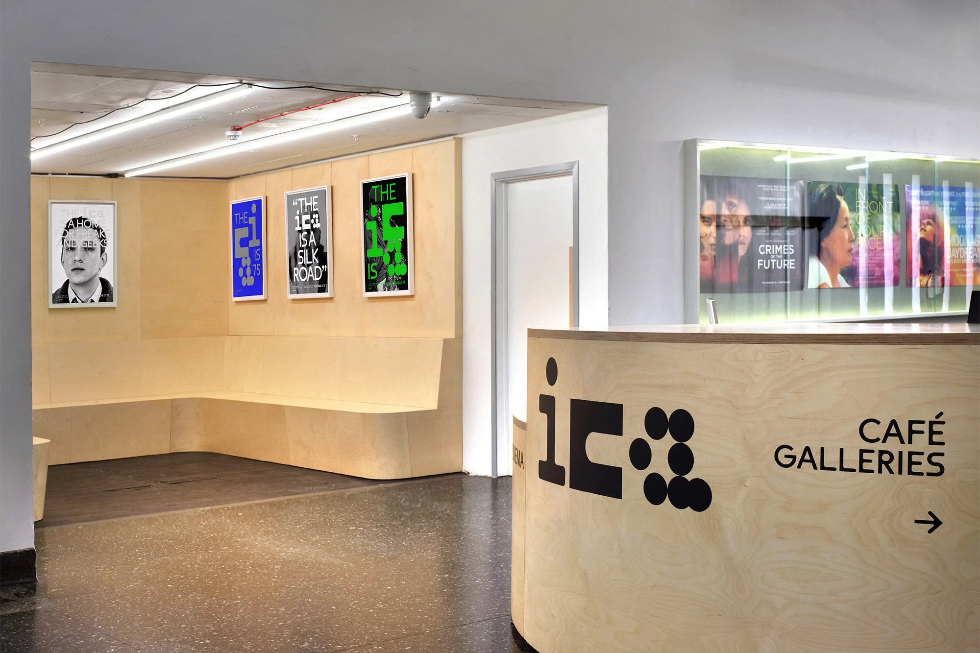 Art gallery reception area with modern blonde wood counter, black logo, graphic art posters, and interior bench seating.