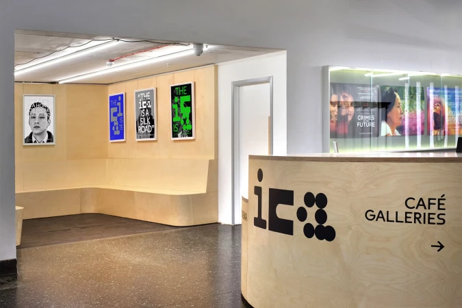 Art gallery reception area with modern blonde wood counter, black logo, graphic art posters, and interior bench seating.