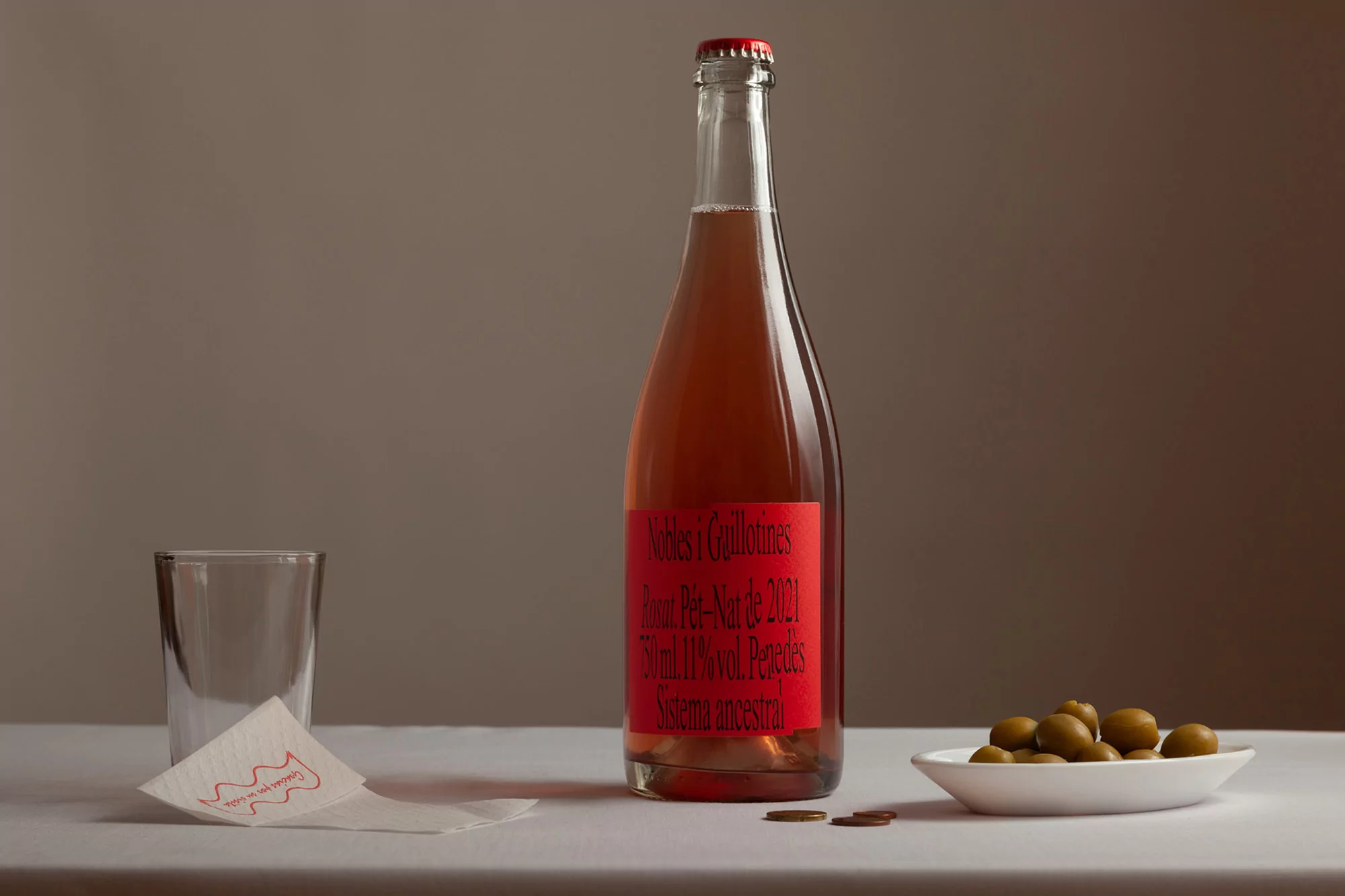 Bottle of rosé wine with a red label sits on a white table with a glass, olives, coins, and a napkin against a neutral backdrop.